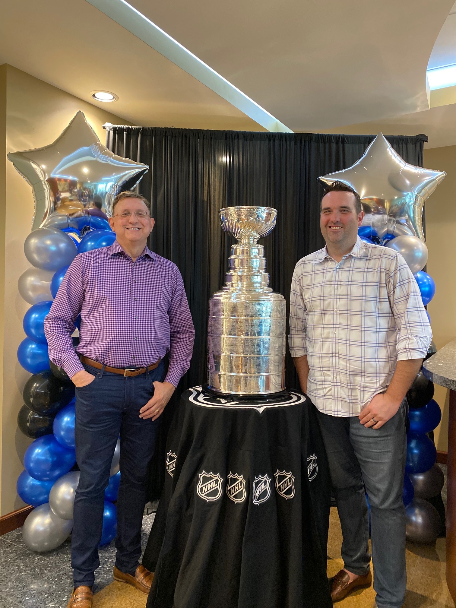 Bob Ritchie and Jon Ritchie standing next to the Stanley Cup with balloons in the background in the color scheme of the Tampa Bay Lightning